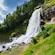 A large waterfall cascading down moss covered rocks surrounded by trees and mountains
